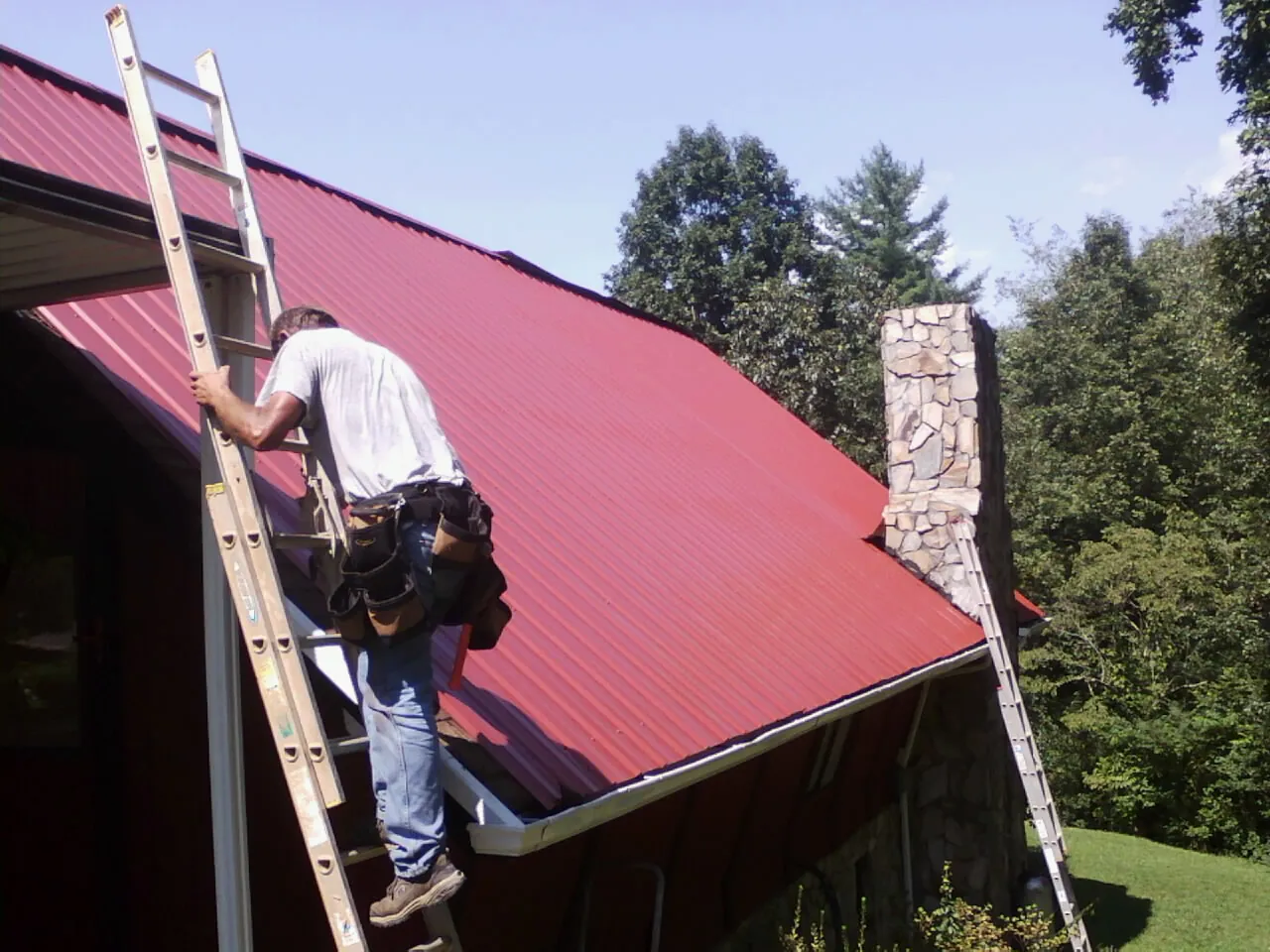 Roofer on a ladder beside a red metal roof and stone chimney — Soffit & Fascia Repair by Roofing Brothers in Saint Michaels